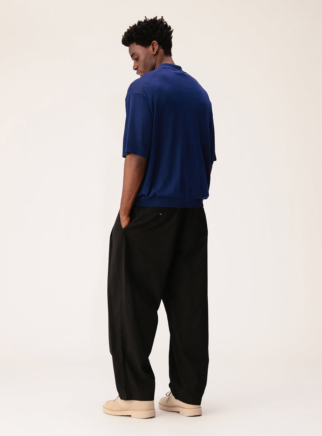 A man faces away with hands in pockets, wearing a relaxed fit blue crepuscule Short Sleeve Polo, wide black pants, and light tan shoes against a plain light background.