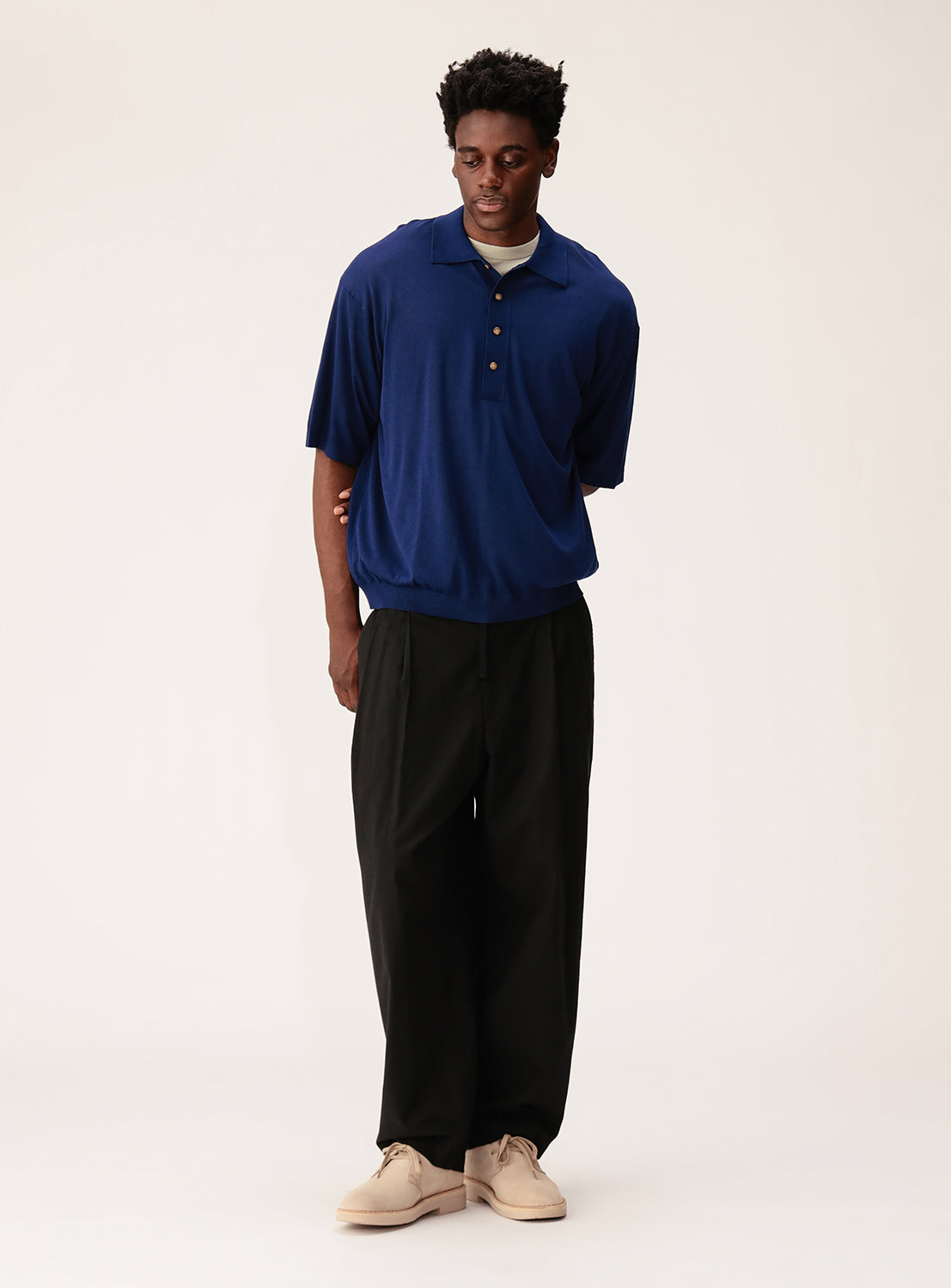 A man stands against a plain light background, wearing the crepuscule Short Sleeve Polo in blue with a relaxed fit, paired with black trousers and beige shoes. He looks slightly downward, one arm behind his back.
