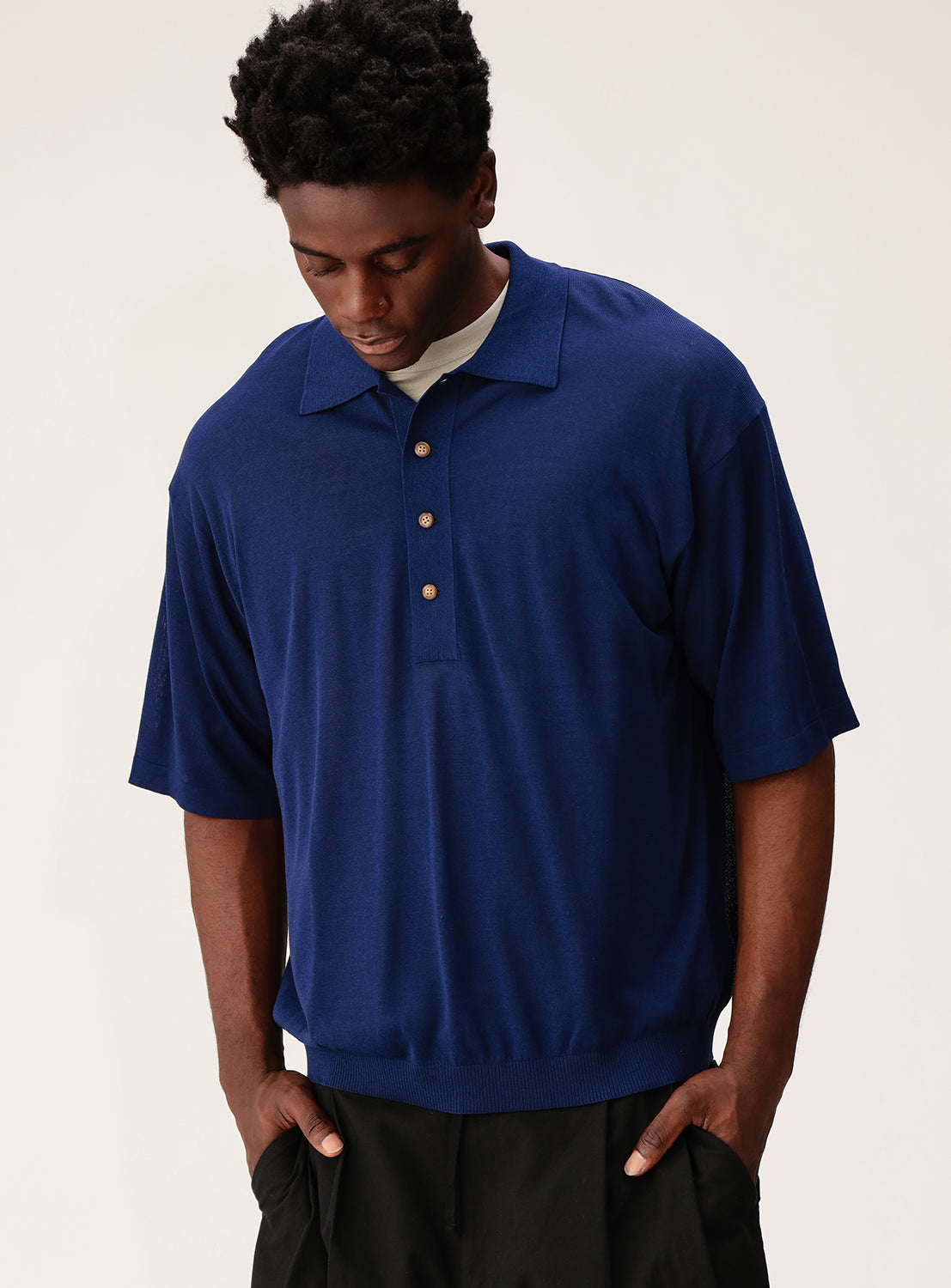 A man wears the crepuscule Short Sleeve Polo in dark blue, featuring a relaxed fit, subtle sheen, and three buttons, paired with black pants. He stands facing slightly downward with hands in pockets against a light background.