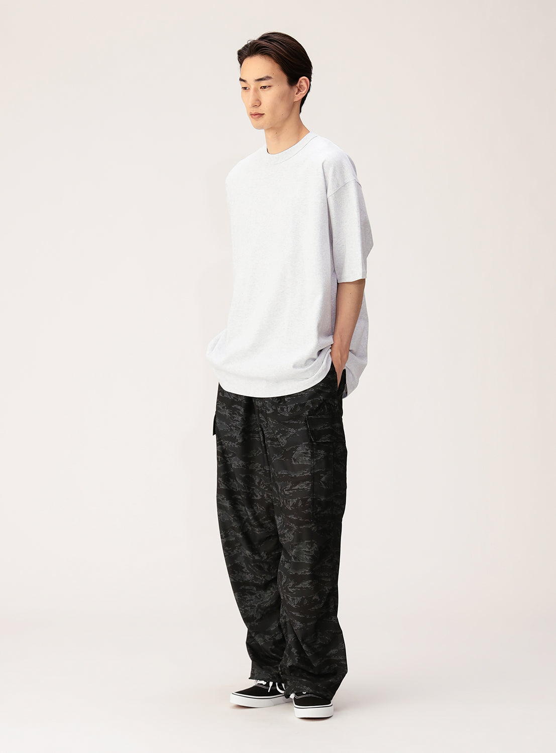 A person stands against a plain backdrop wearing the NEW T Heavyweight Blank T-Shirt, crafted from open-end yarn cotton, paired with baggy dark camouflage pants and black sneakers. With hands in pockets and a relaxed expression, this look perfectly embodies unisex style.
