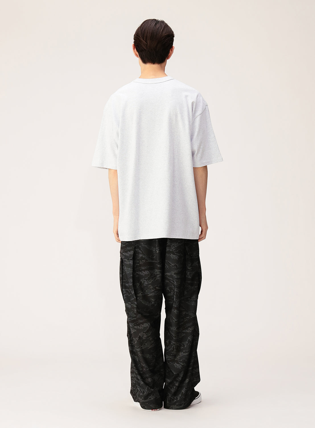A person stands facing away, wearing the NEW T Heavyweight Blank T-Shirt crafted from open-end yarn cotton and baggy black patterned pants against a plain light background, showcasing its unisex wear appeal.
