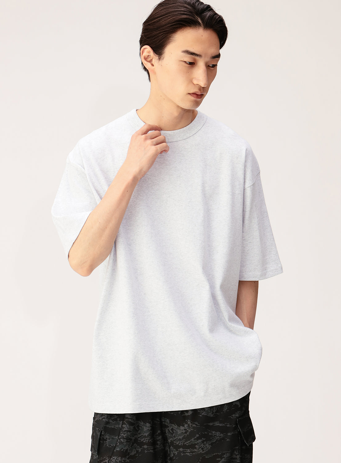 A young man with dark hair wears the NEW T Heavyweight Blank T-Shirt, crafted from open-end yarn cotton, paired with dark patterned pants. Standing against a plain background and looking down with one hand near his collar, he showcases the versatile and relaxed appeal of this unisex tee.