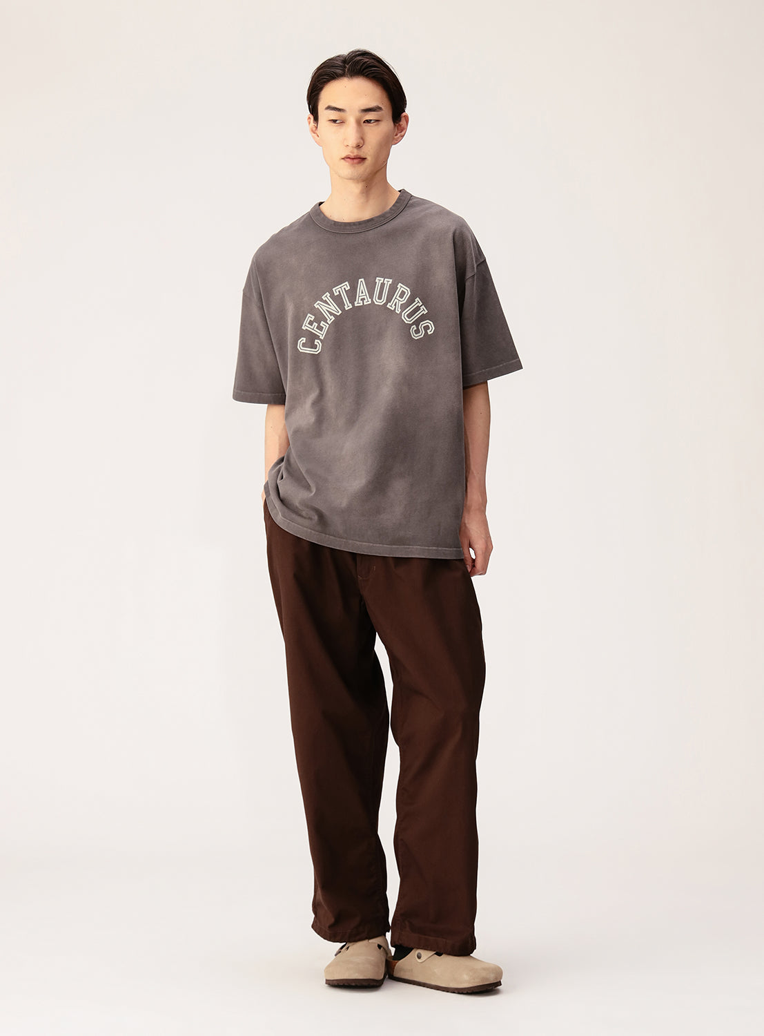 A young man with straight dark hair wears a BEAMS Souvenir T-Shirt and loose brown pants with beige slip-on shoes, standing in a relaxed pose against a plain light background.
