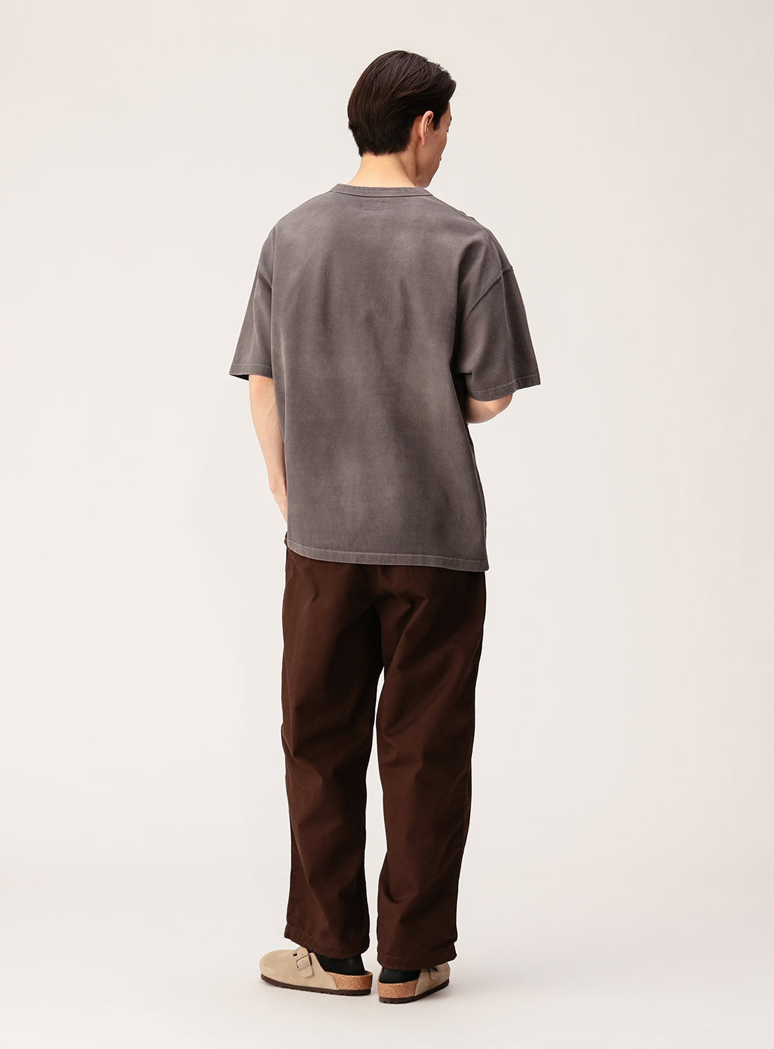 A person stands with their back to the camera, wearing the BEAMS Souvenir T-Shirt in loose gray cotton jersey, paired with brown pants and beige shoes against a simple, light background.
