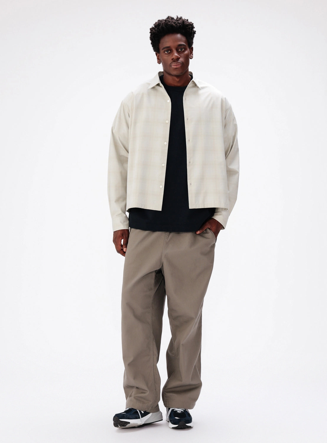 A man wearing a BEAMS Shadow Check Shirt in cream over a black t-shirt, paired with wide-fit taupe trousers and sneakers.