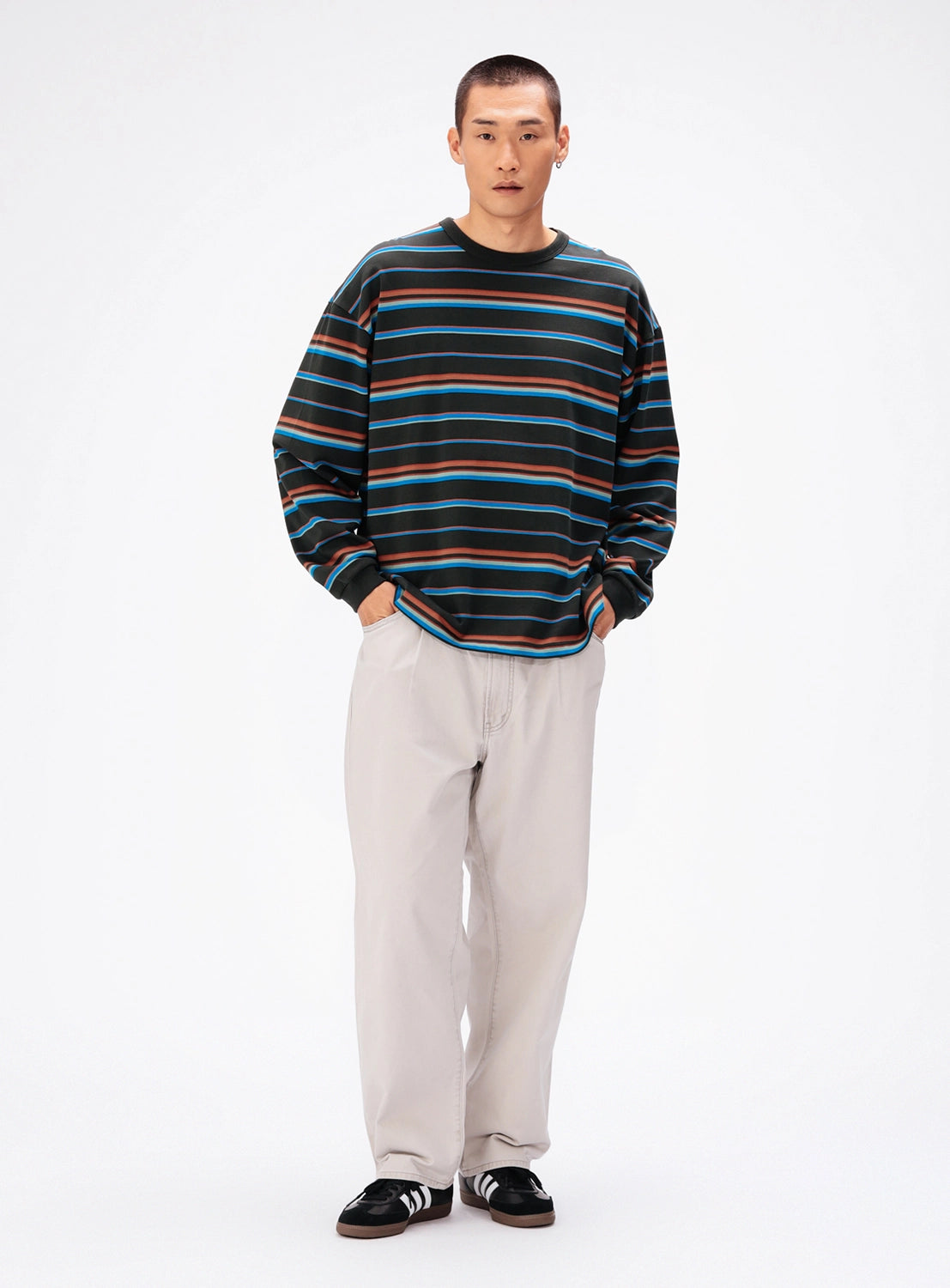 A man wears a BEAMS Loose Fit Border Long Sleeve T-Shirt in black, blue, and brown, paired with light-colored wide fit trousers and black sneakers.