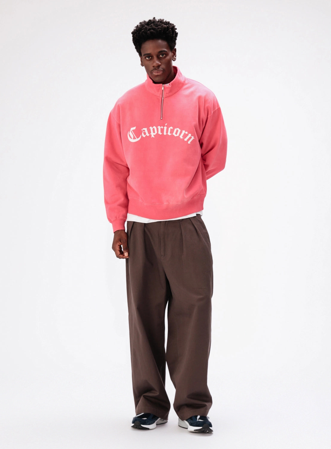 A person wears a BEAMS Faded Souvenir Half Zip Pullover in pink with Capricorn embroidered on the chest, paired with brown trousers and dark sneakers.