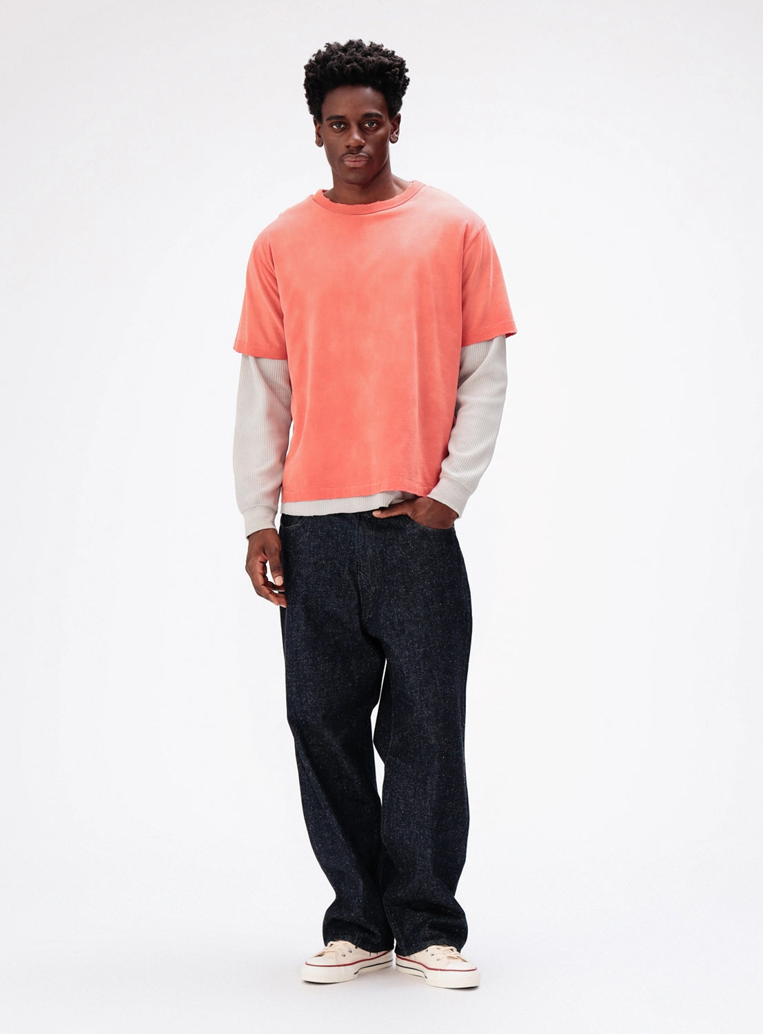 A person wearing a BEAMS Vintage Layered Fade Long-Sleeve T-Shirt in coral over a gray long-sleeved shirt, paired with dark denim jeans and canvas sneakers.