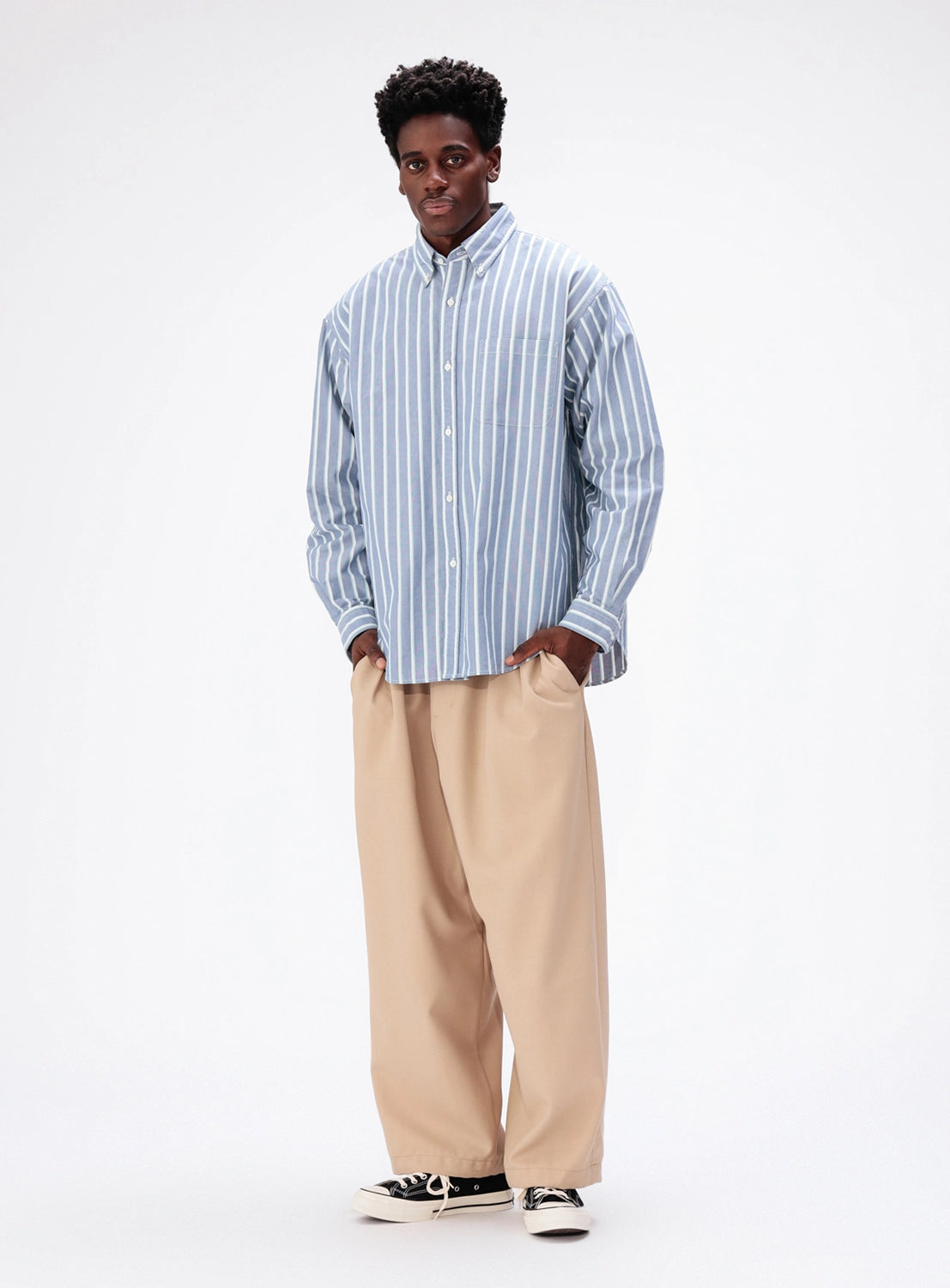A person wearing a blue and white striped BEAMS Heavy Oxford Button-Down tucked into loose-fitting tan trousers and black sneakers.