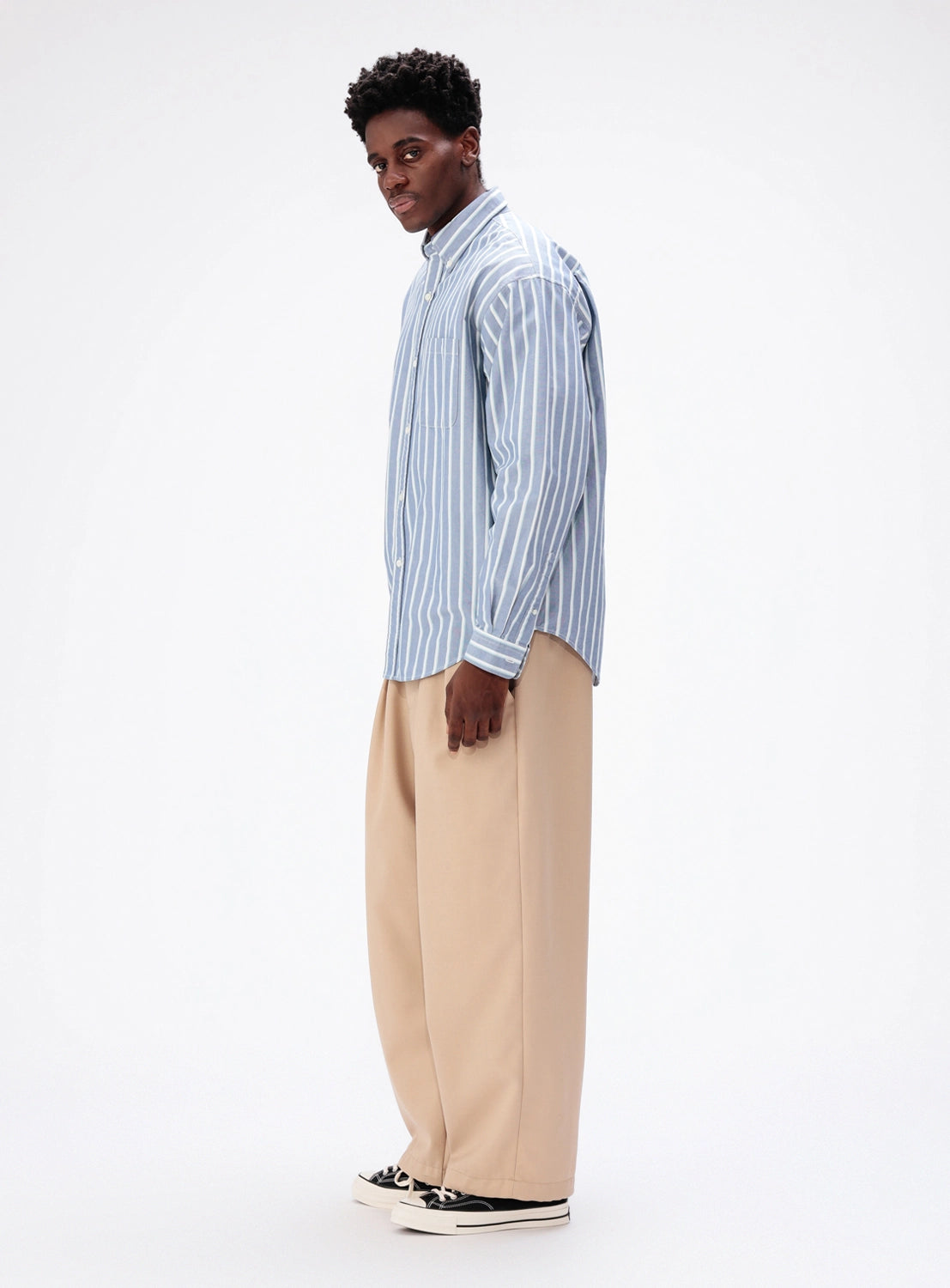 A man standing sideways wearing a BEAMS Heavy Oxford Button-Down shirt in blue and white stripes with loose-fitting tan trousers.
