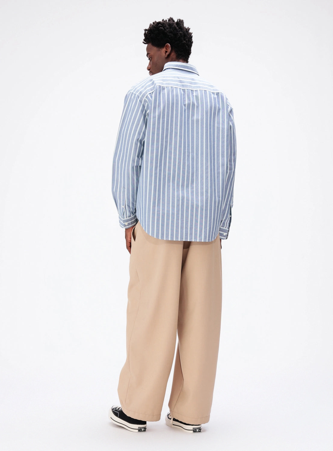 A person seen from behind wears a blue-and-white striped BEAMS Heavy Oxford Button-Down Shirt tucked into wide-leg tan trousers with black sneakers.