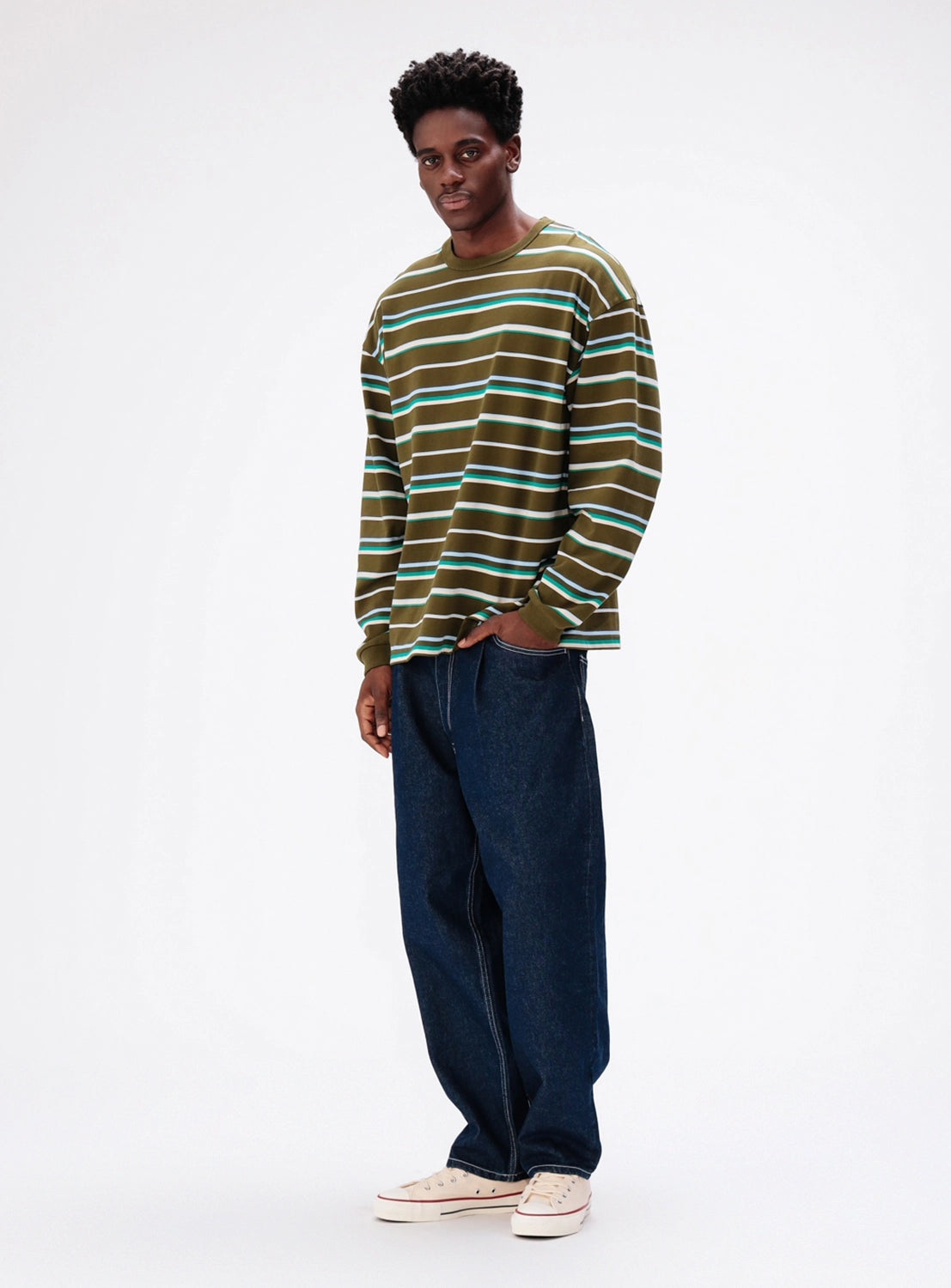 A man wears a BEAMS Loose Fit Border Long Sleeve T-shirt in olive, white, and teal, paired with wide-fit dark denim jeans and canvas sneakers.