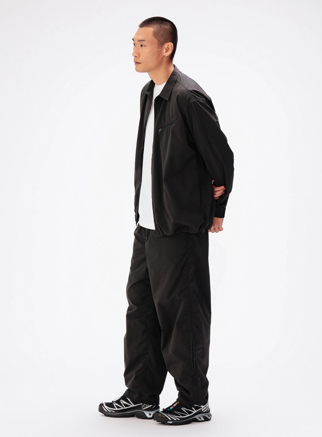 A man wears a black BEAMS Tech Shirt over a white t-shirt, paired with matching black trousers and black sneakers.