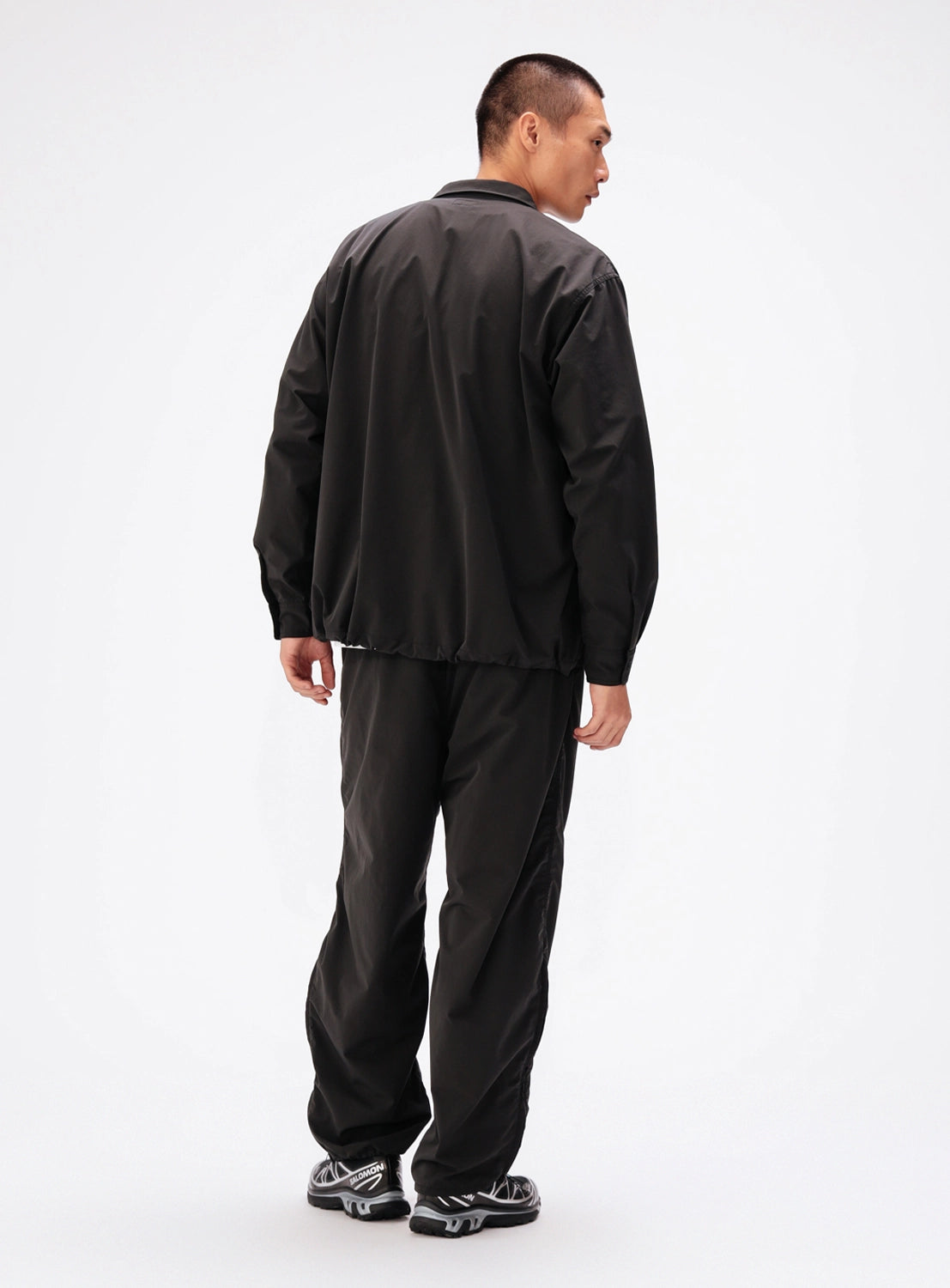 A person seen from behind, wearing a black long-sleeved BEAMS Tech Shirt with a collar, matching black pants, and grey running shoes.