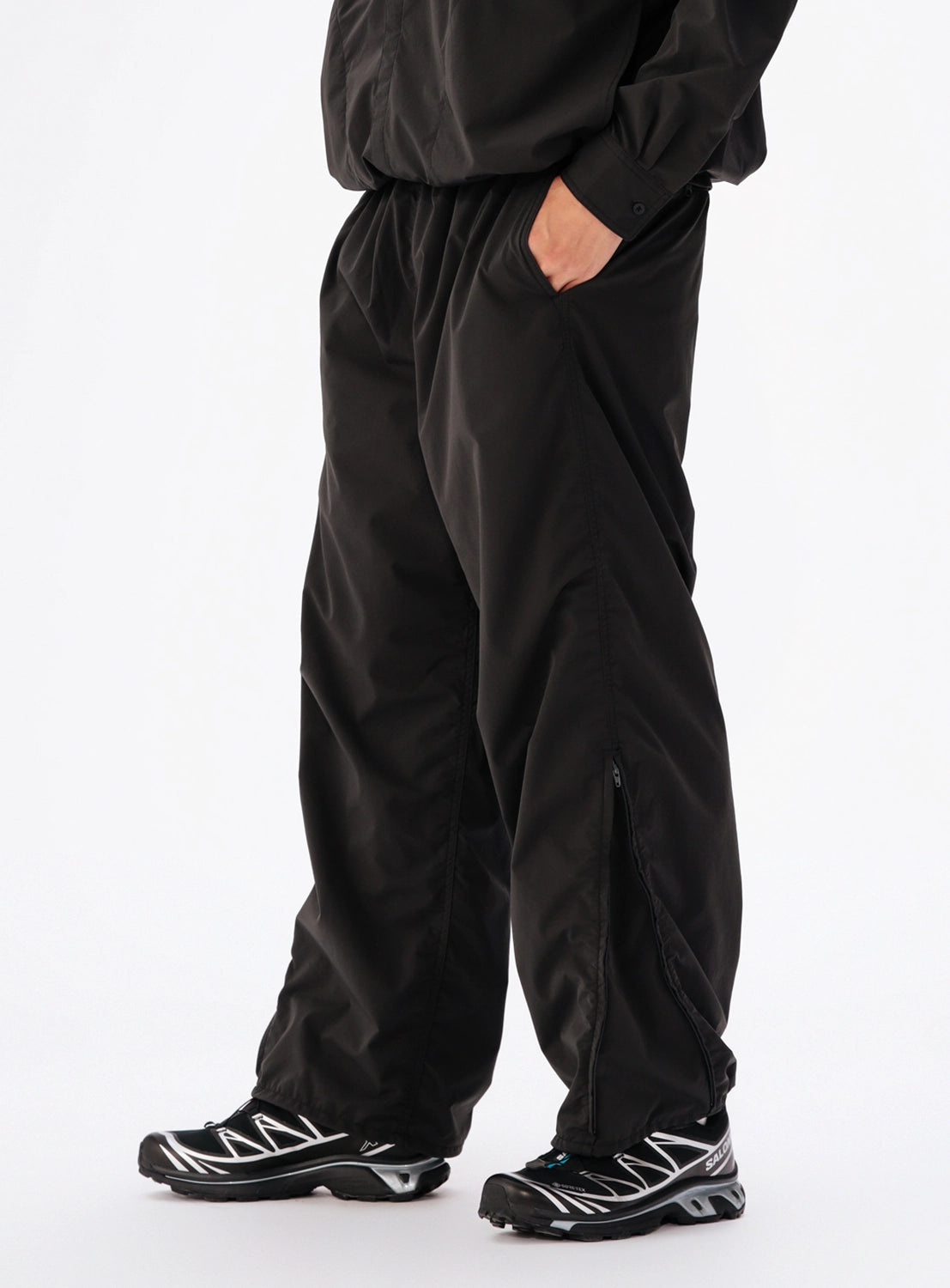 A person wearing BEAMS Technical 2-Way Adjustable Easy Pants, which are wide, straight, water-repellent trousers with zippered hems, paired with athletic sneakers and a hand tucked into a side pocket.