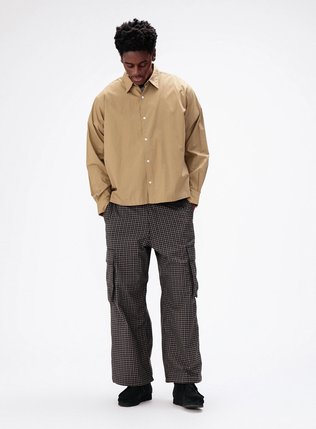 A person wearing a tan long-sleeved button-down shirt, dark checked BEAMS Pattern 6-Pocket Pants in a relaxed fit with six pockets, and dark shoes.