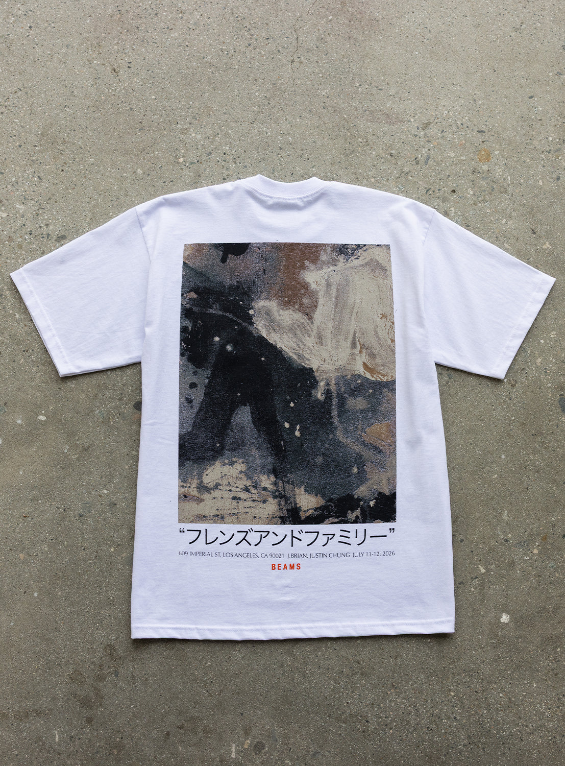 The J. BRIAN x BEAMS TEE by BEAMS AMERICA is a limited edition white T-shirt, laid flat on a speckled surface, showcasing original abstract artwork on the back with Japanese text, English phrases, and a bold red “BEAMS” logo near the bottom hem—a standout piece perfect for collectors and style enthusiasts alike.