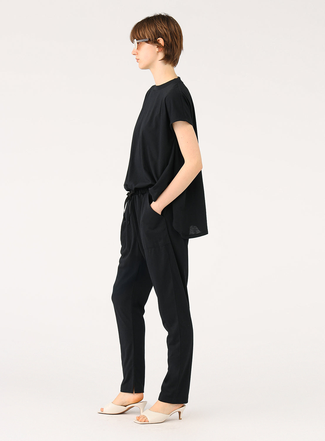 Side profile of a model wearing a black jumpsuit, highlighting the cinched waist and the tapered legs.
