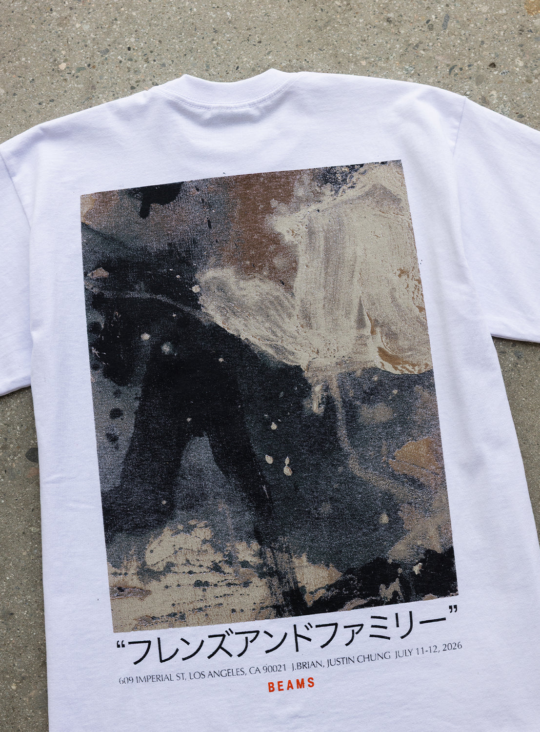 J. BRIAN x BEAMS TEE by BEAMS AMERICA—a limited edition white t-shirt featuring a textured, abstract art print on the back. Below the original artwork are Japanese characters, the phrase “FRENDS AND FAMILY,” and event details in English. The shirt is displayed flat against a speckled gray surface.
