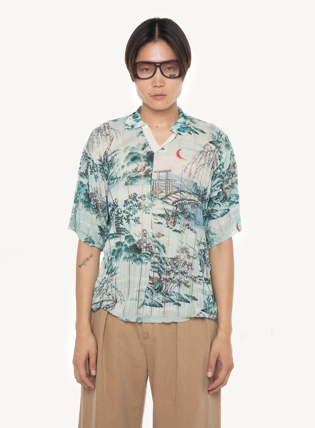 A person stands facing forward against a plain white background wearing sunglasses, tan pants, and the DAIRIKU Japanese Pattern Aloha Transfer Print Polo Knit in light blue with nature-inspired prints.