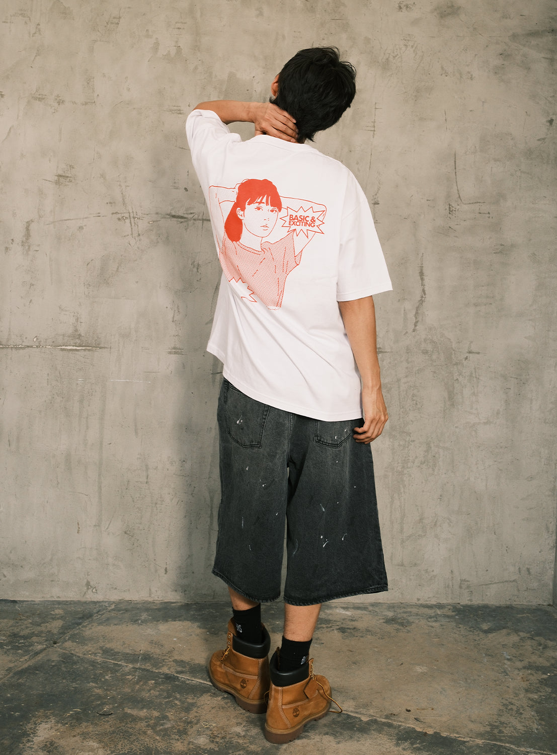 A person stands with their back to the camera, hand on neck, wearing a white BEAMS T “BEAMS 50th Anniversary x KYNE T-Shirt” made from heavyweight jersey cotton and featuring a red line art portrait. They pair it with loose black shorts, tan boots, and black socks against a textured gray wall—celebrating the BEAMS 50th anniversary with effortless style.