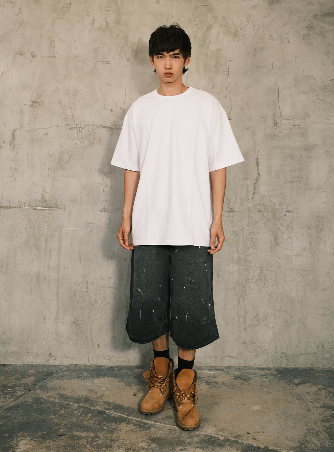 A young man stands indoors against a textured gray wall, wearing a loose heavyweight jersey cotton BEAMS 50th Anniversary x KYNE T-Shirt by BEAMS T in white, dark baggy shorts with subtle paint marks, and tan work boots. He looks at the camera with a neutral expression.