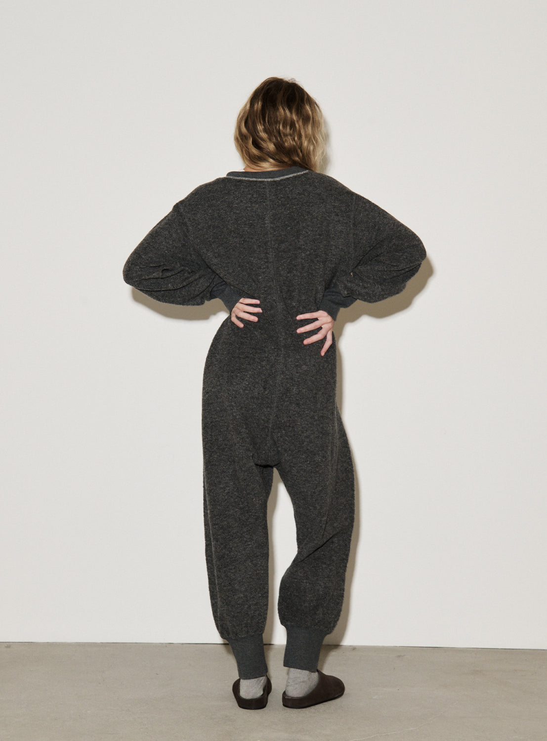 A person stands facing a white wall with hands on hips, wearing THE BASIC Knit Jumpsuit in dark gray—a relaxed, essential wardrobe staple—paired with gray socks and slippers. Their shoulder-length hair is wavy.