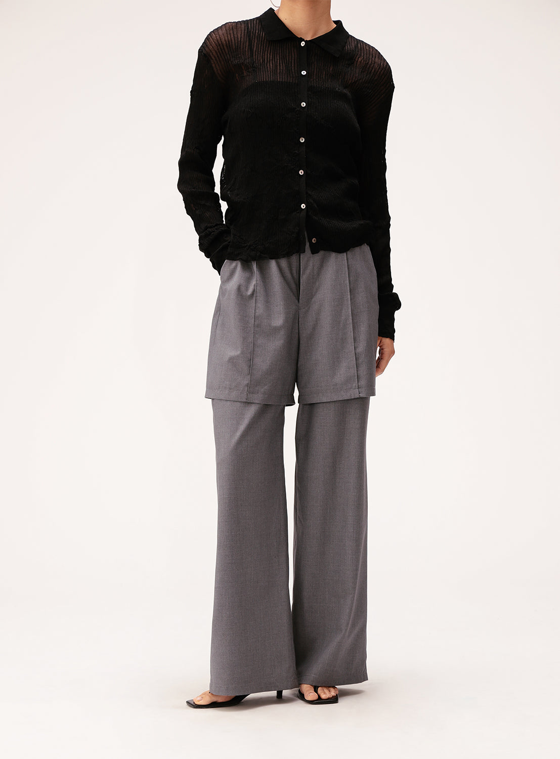 A person is wearing a sheer black button-up shirt, loose gray shorts layered over Ray BEAMS’ Half Layered Wide-Leg Trousers crafted from lightweight suiting fabric, and black open-toe shoes, standing against a plain white background.