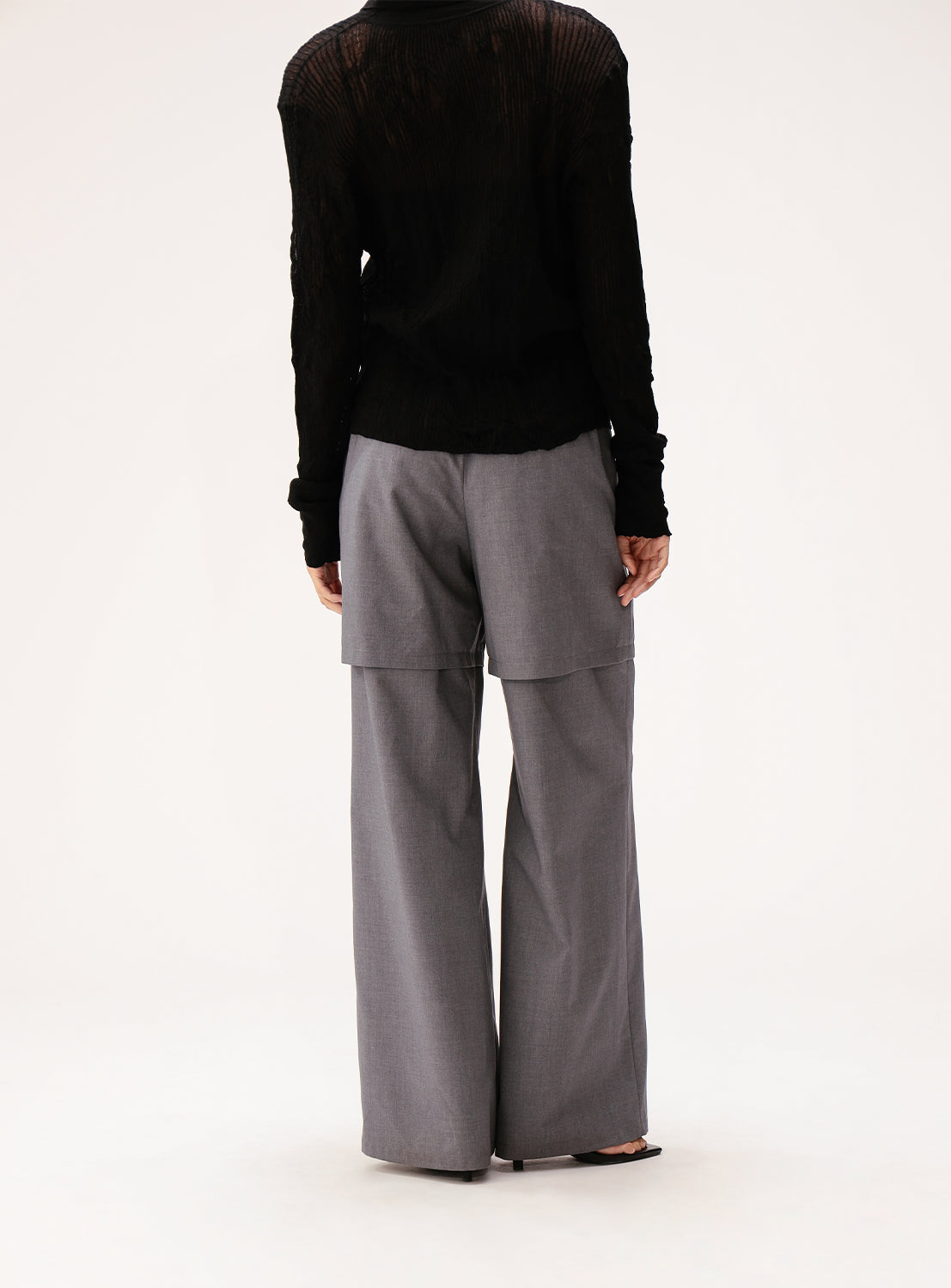 A person stands facing away, wearing a sheer black long-sleeve top paired with Ray BEAMS' Half Layered Wide-Leg Trousers in gray, featuring an overlapping panel detail at the back. The background is plain and light-colored.