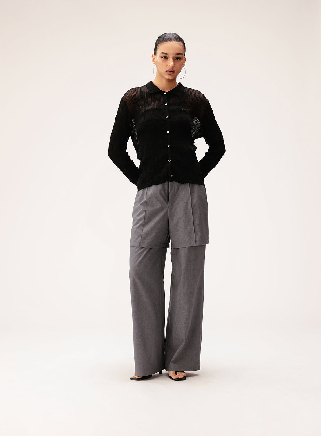A person stands confidently with hands in pockets, wearing the Ray BEAMS Half Layered Wide-Leg Trousers—loose gray trousers crafted from lightweight suiting fabric—paired with a black button-up top, posed against a plain light background.