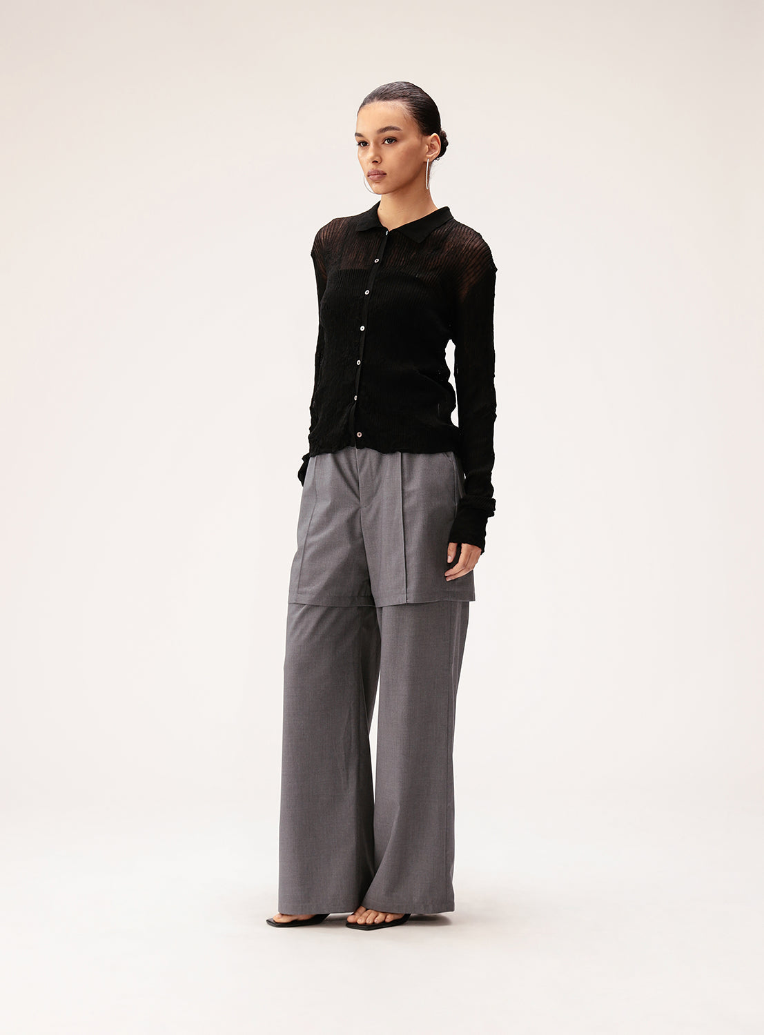 A woman stands against a plain background wearing Ray BEAMS' Half Layered Wide-Leg Trousers in gray, paired with a black button-up top. Her hair is pulled back and she gazes slightly to the side with a neutral expression, highlighting the wide, straight silhouette of the trousers.