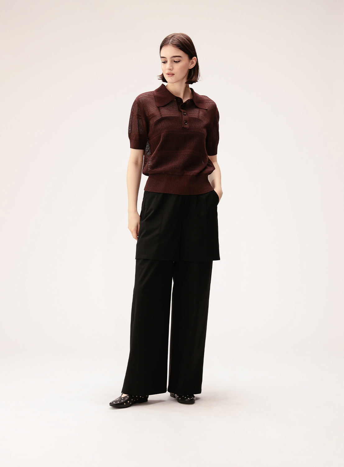 A woman stands against a plain background, wearing a short-sleeved maroon knit top, black shoes with small studs, and Ray BEAMS' Half Layered Wide-Leg Trousers—crafted from lightweight suiting fabric in a wide, straight silhouette. She looks to her left with her hands in her pockets.