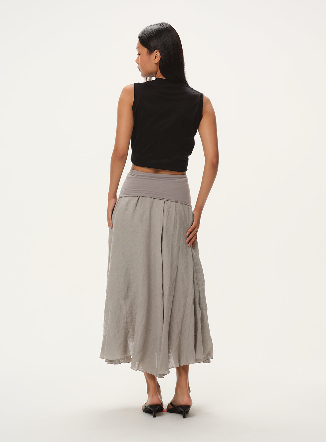 A female model from behind, wearing a light grey, full midi skirt with a black sleeveless crop top.