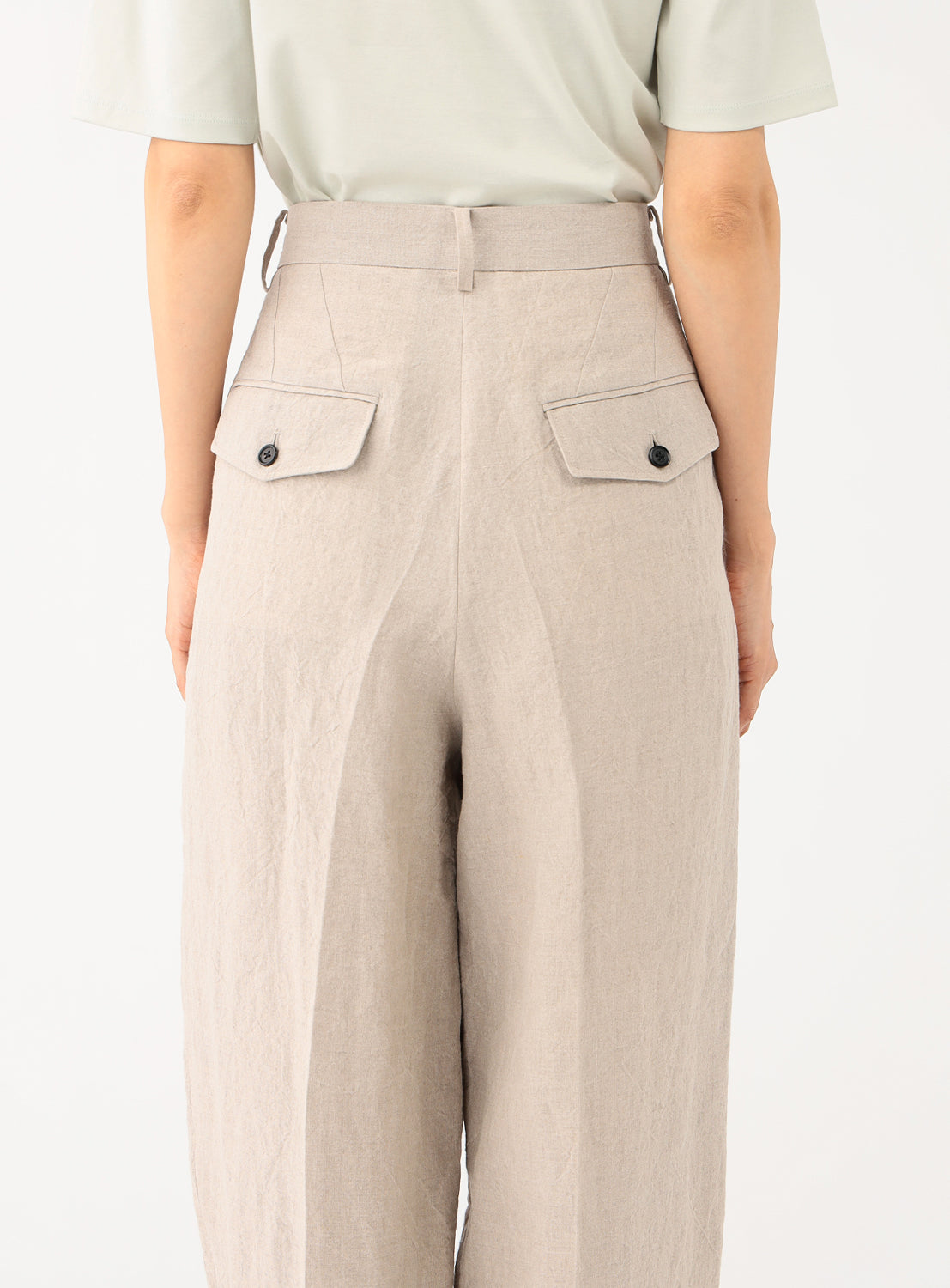 Back view of a model in the light beige pants, highlighting the pleats and buttoned back pockets.