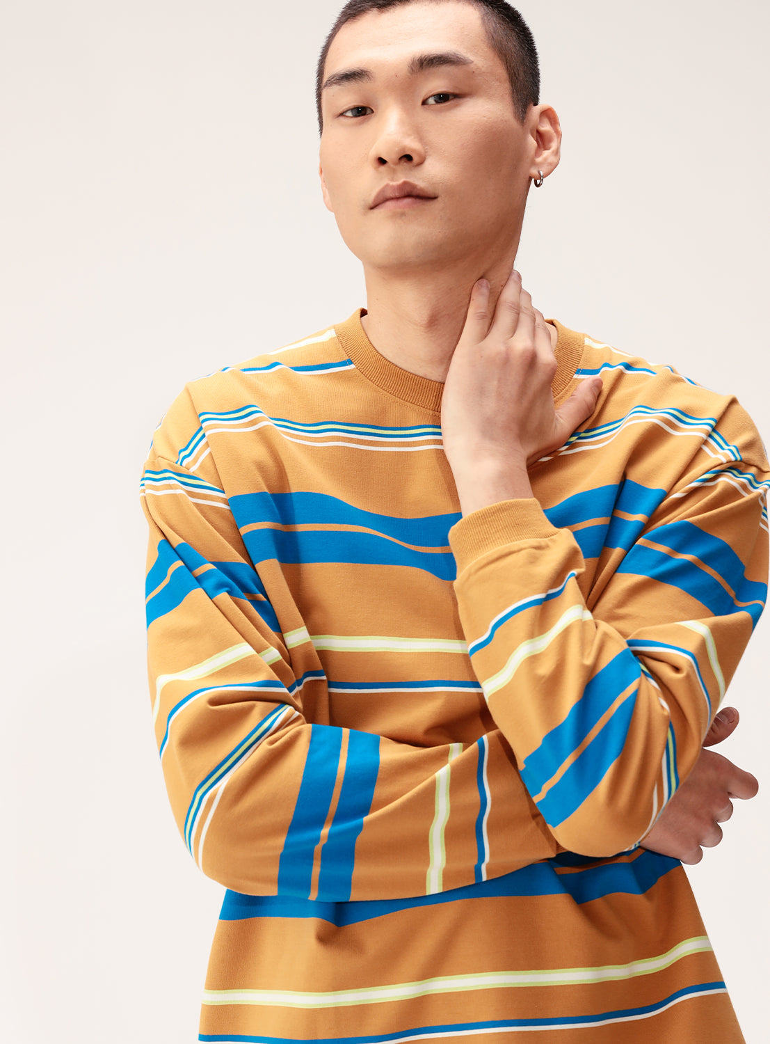 A person with short hair wears the BEAMS Multi-Stripe Crew Neck Wide Fit Sweatshirt in mustard yellow, blue, and white, standing against a light background, looking at the camera with one hand on their neck and the other arm crossed.