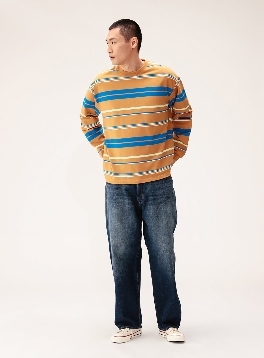 A man stands against a light background, wearing the BEAMS Multi-Stripe Crew Neck Wide Fit Sweatshirt in mustard yellow with blue and white stripes, paired with blue jeans and white sneakers, looking left with his hands behind his back.