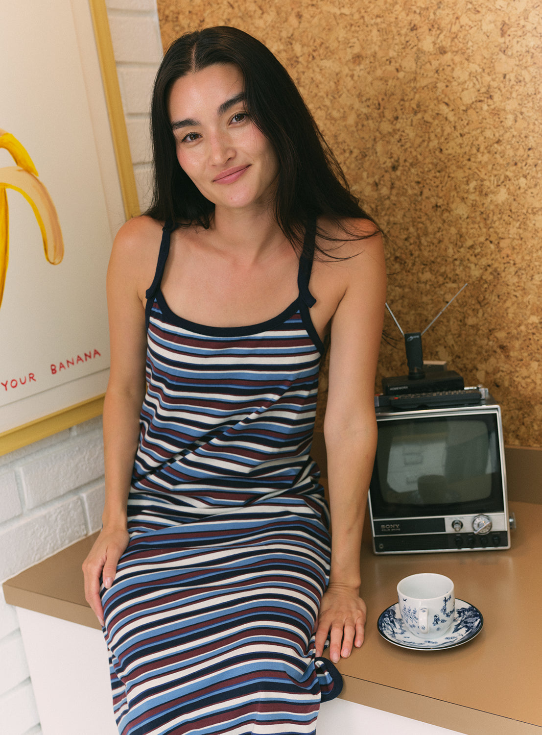 Model wearing a blue, burgundy, and white striped dress with navy blue straps and a navy blue trim, sitting on a kitchen counter.