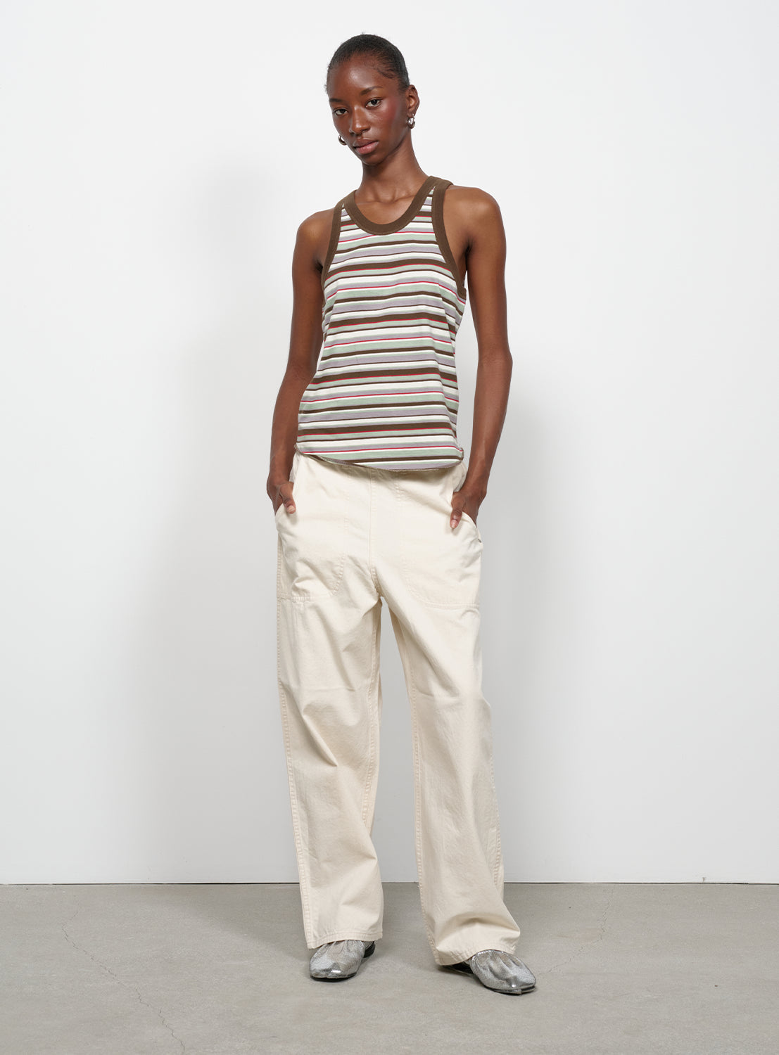 Full body view of a model wearing a multi-colored striped tank top with brown trim and white pants. 