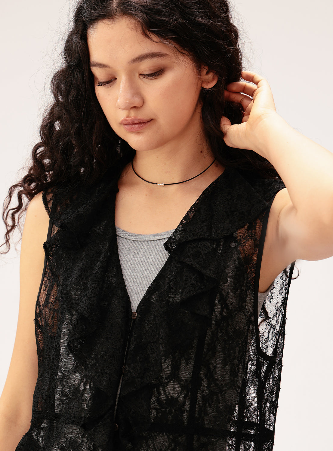 A woman with long curly hair wears a sheer black lace vest over a gray top, accessorized with the Ray BEAMS Silver Top Leather Choker. She looks down softly, touching her hair with one hand.