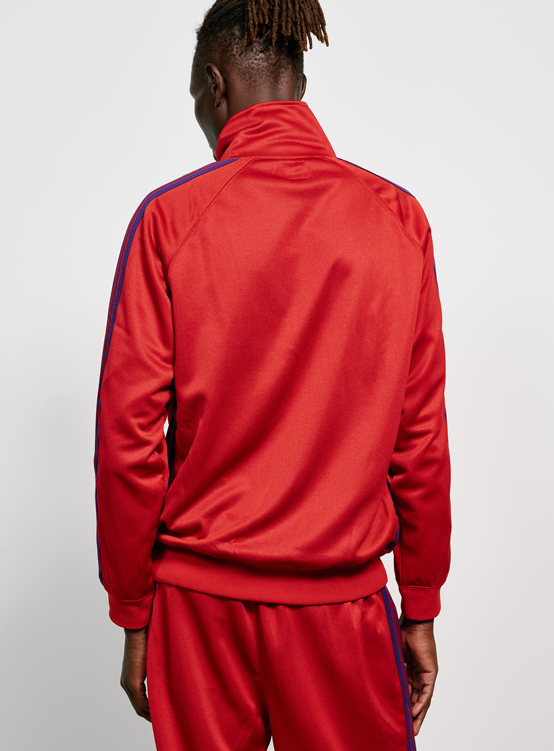 Backside of a model wearing a red track jacket with blue/purple stripes, front pockets with zippers and an embroidered purple butterfly. 