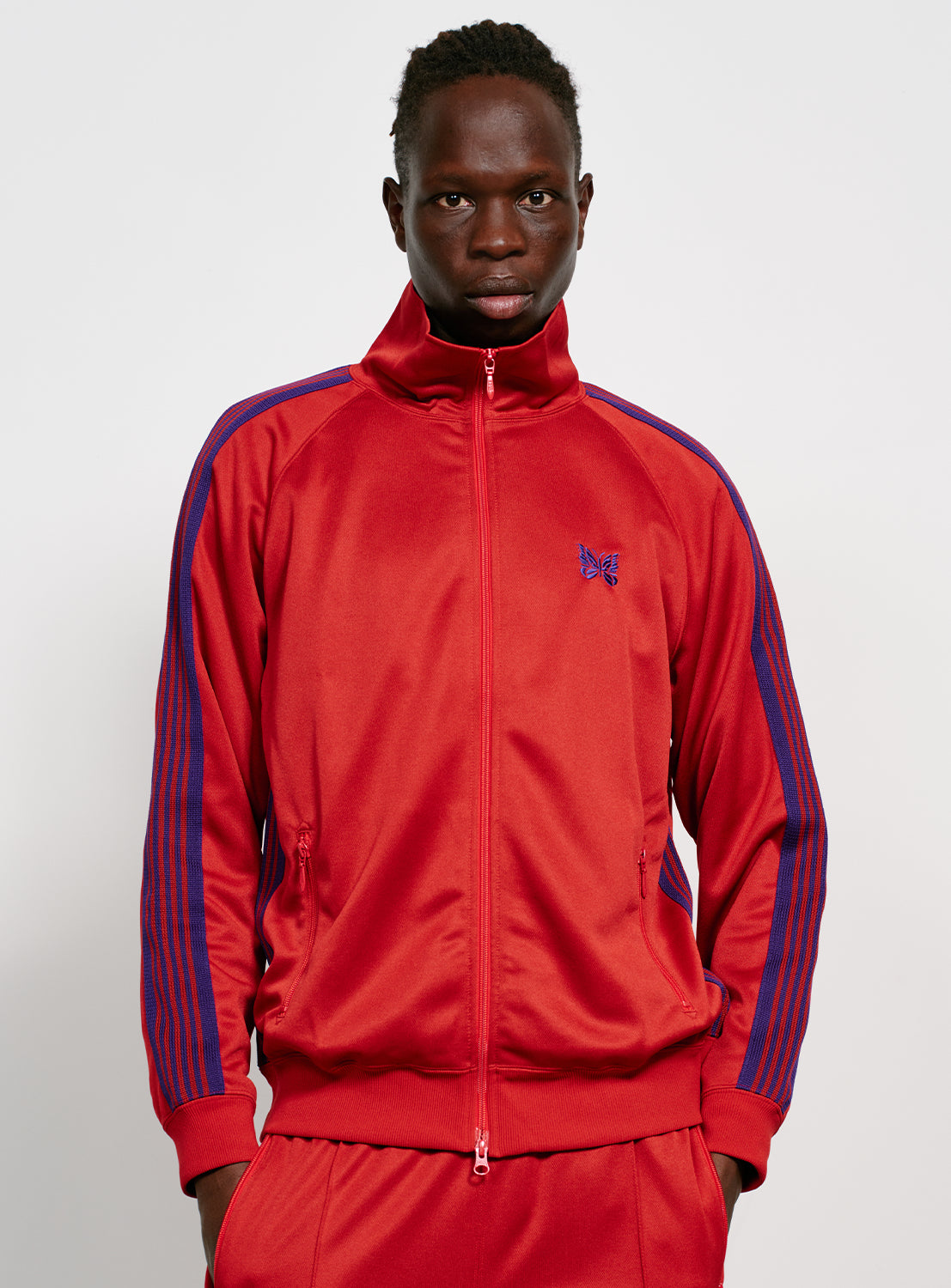 Model wearing a red track jacket with blue/purple stripes, front pockets with zippers and an embroidered purple butterfly. 