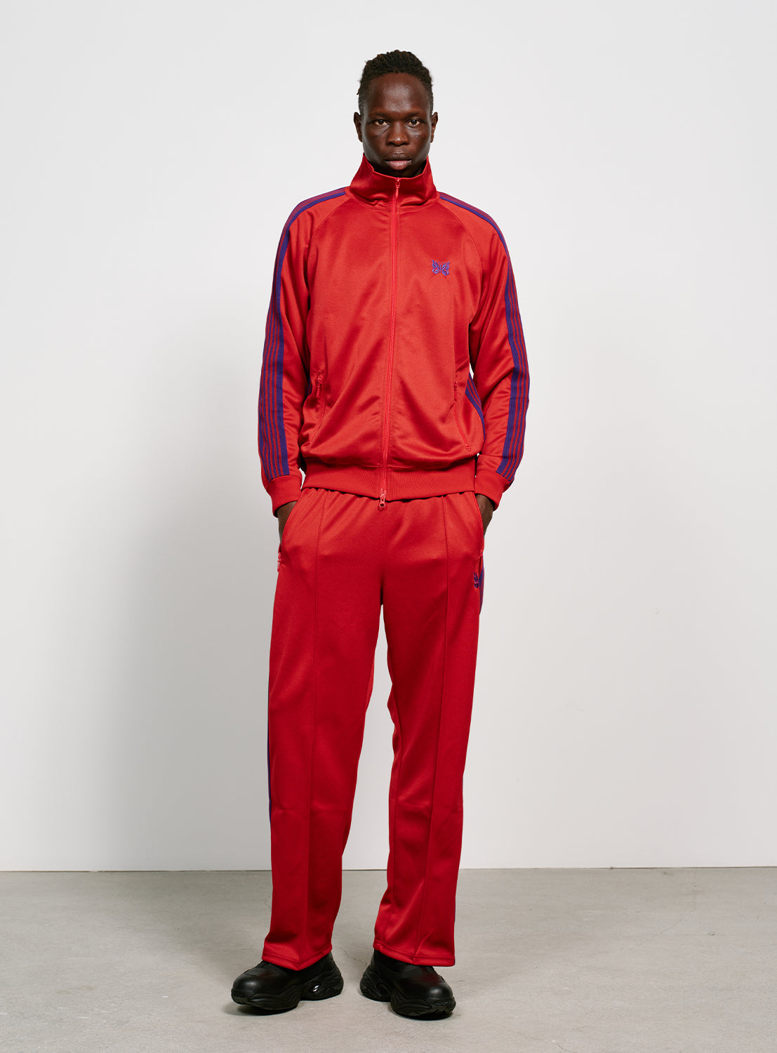 Model wearing a red track jacket with blue/purple stripes with the matching pants.