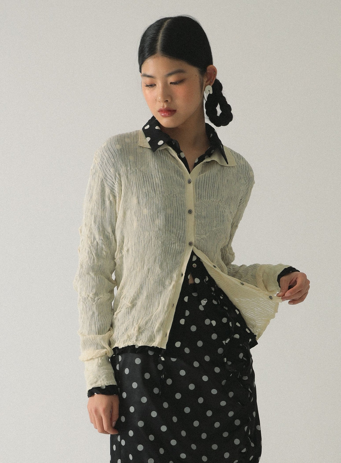 A woman with braided hair wears the Ray BEAMS Sheer Rib Knit Polo Cardigan over a black polka-dot dress with a matching collar, standing against a plain background and looking down and to the side.