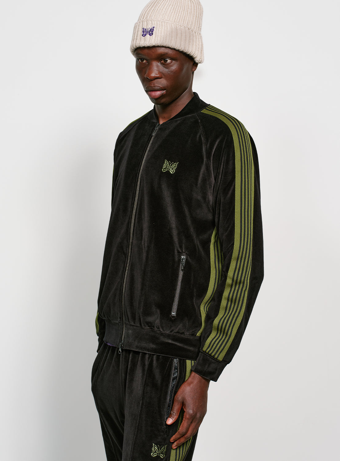 Model wearing a black velour jacket with bright green stripes on the sleeves and side, with front zipper pockets and a green embroidered butterfly on the chest. 