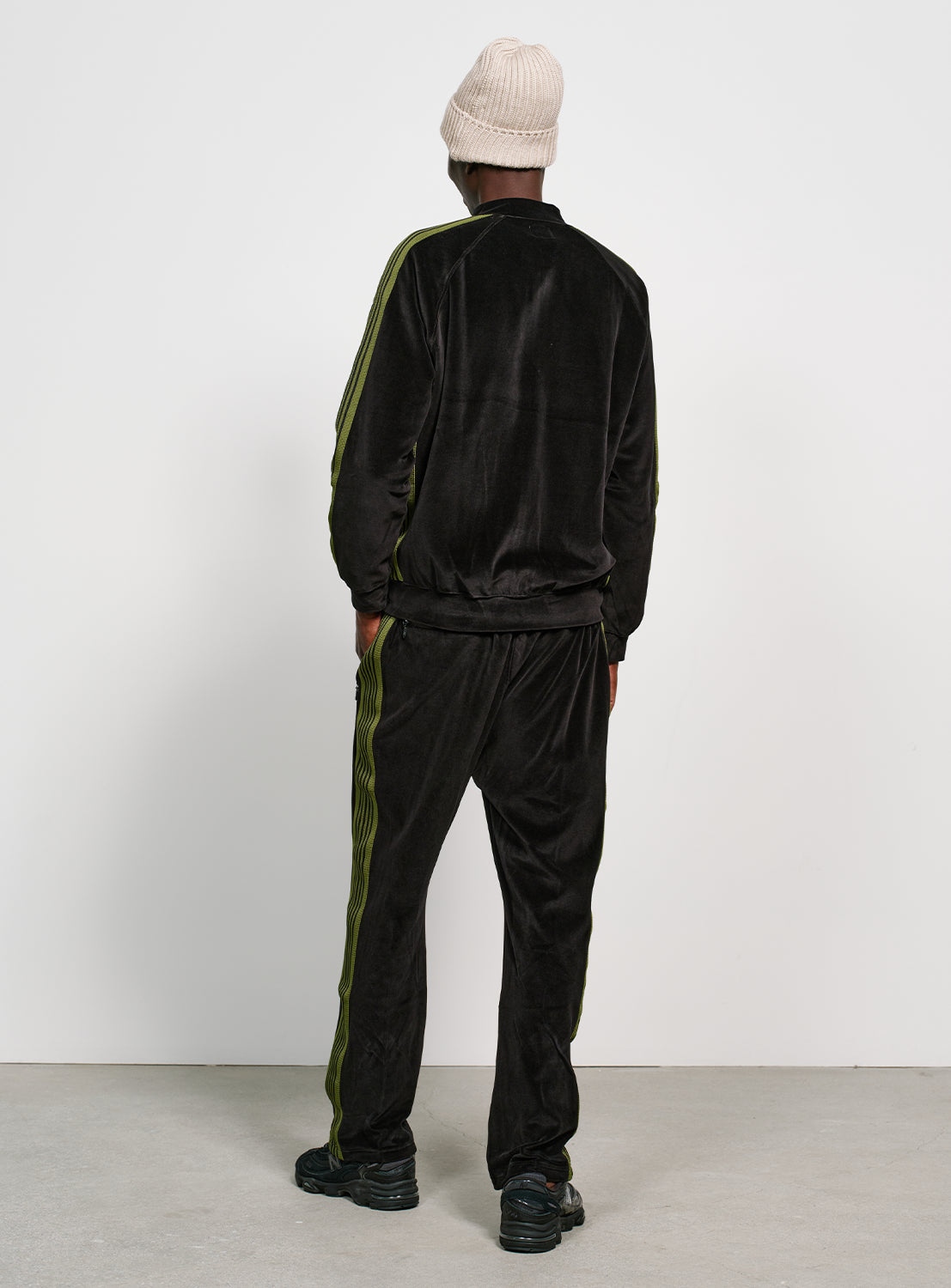 Backside of a model wearing a black velour jacket with bright green stripes on the sleeves and side, with the matching pants. 