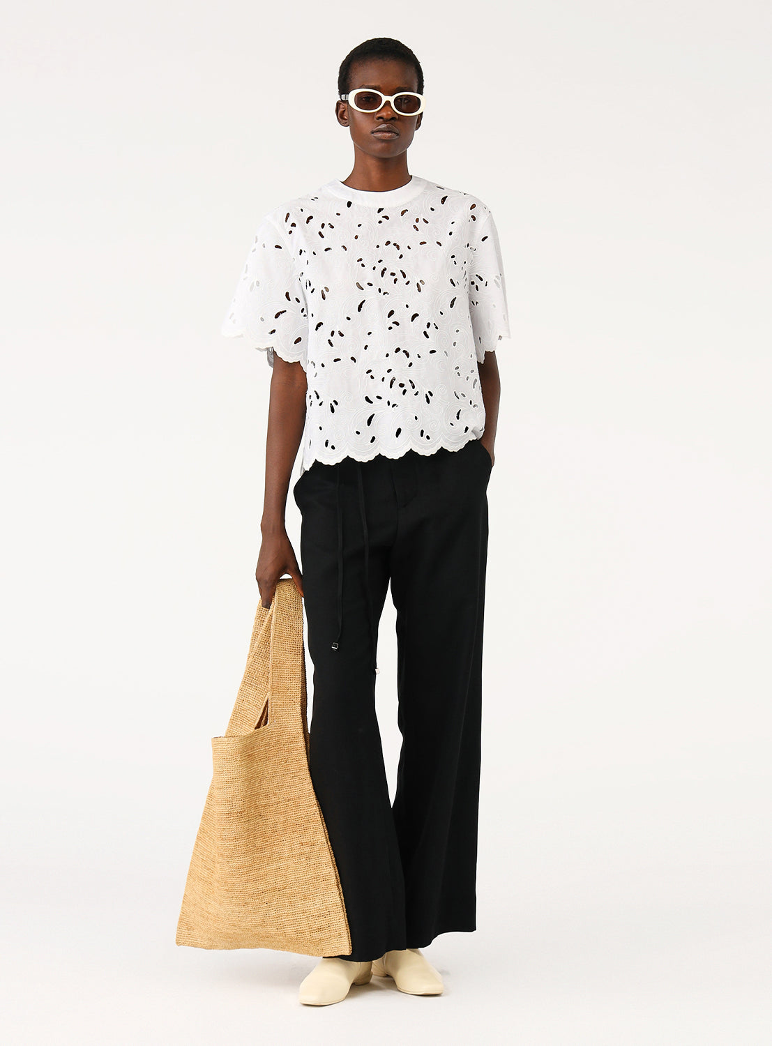 A female model wearing black wide-leg drawstring pants and a white patterned top, holding a paper bag, standing facing forward.