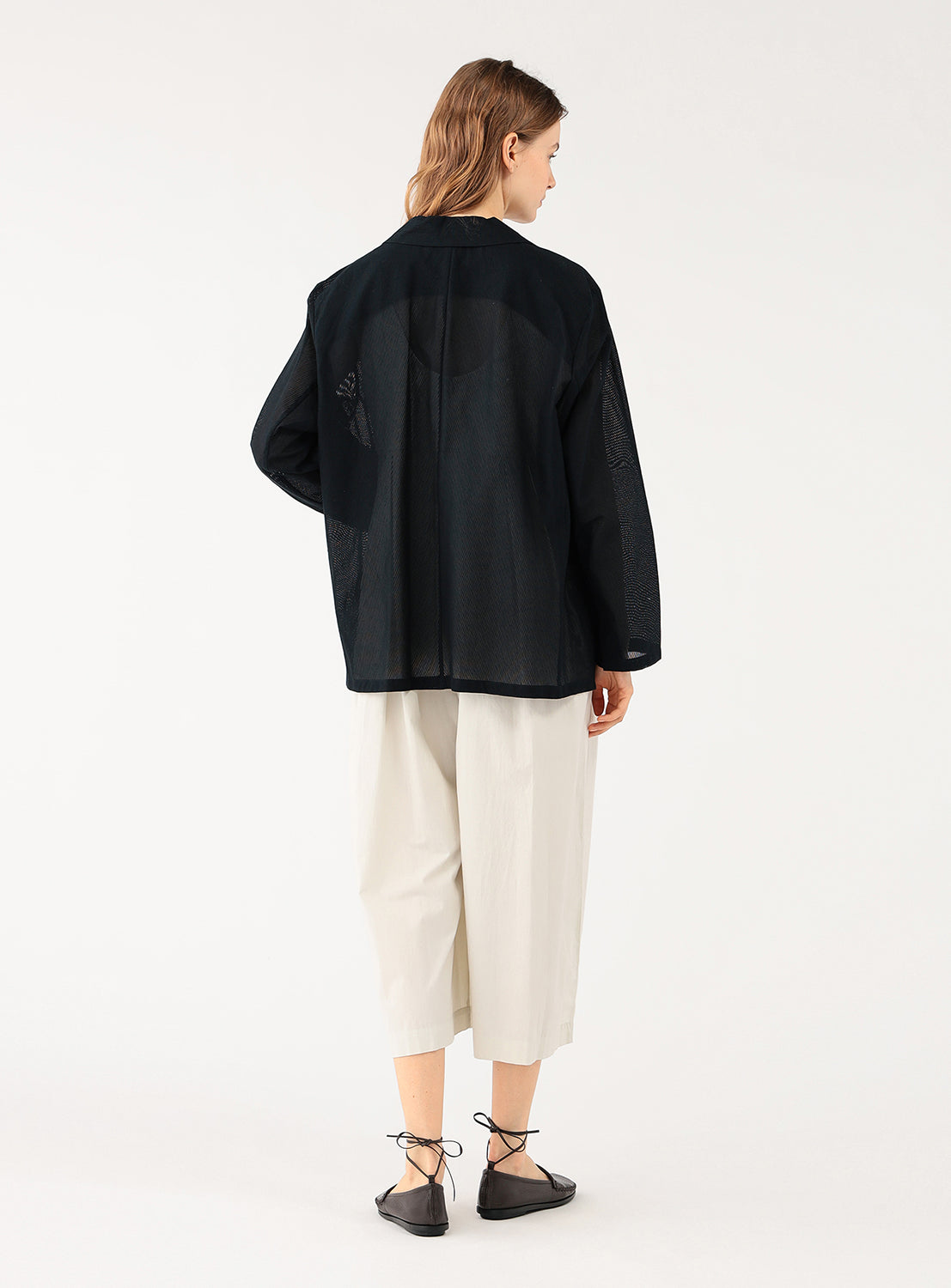 A female model from behind, wearing a dark navy blue open blazer over a light top and cream-colored wide-leg pants.