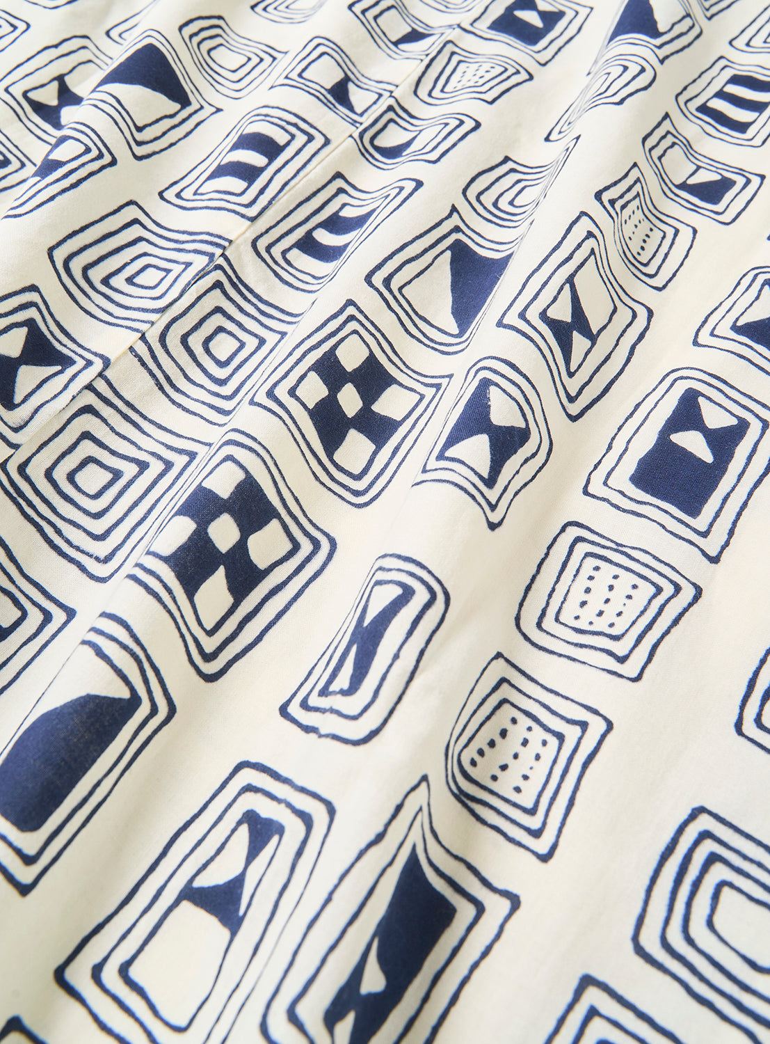The Tutaee Men’s Yukata by Tutaee showcases a white cotton fabric adorned with a repeating pattern of navy blue geometric shapes—including squares, triangles, and abstract motifs. With gentle folds and subtle shadows accentuating its textured design, this traditional Japanese garment is ideal for men seeking comfort and style.