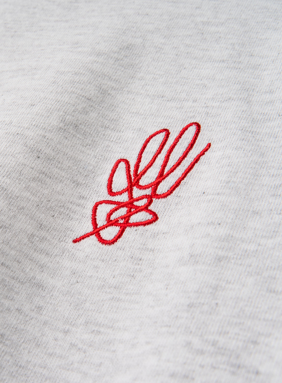 A close-up of the norahi Starskel Hologram T-Shirt in light gray heathered fabric showcases a small, red embroidered abstract design—reminiscent of a hand-drawn motif like a stylized knot or floral flourish—that adds an artistic touch to this relaxed fit graphic tee from norahi.