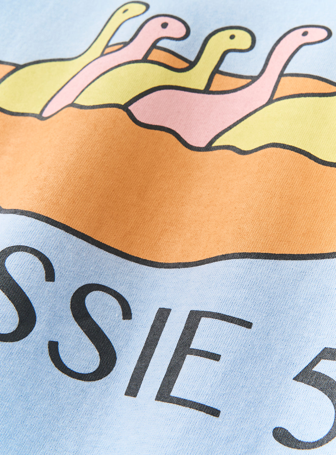 An illustration of four cartoon dinosaurs in pastel hues, evoking a vintage-inspired aesthetic, appears above partially hidden text reading "NESSIE 3" on a light blue background—making the DAISAK Nessie T-Shirt by DAISAK an ideal choice for a playful graphic tee.